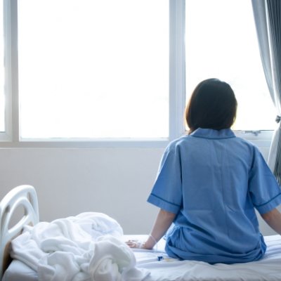 woman in blue shirt sits on a bed and looks out of a window